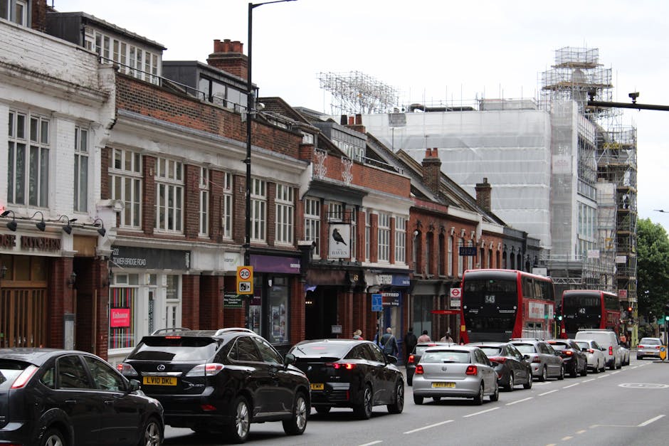 A row of red double-decker buses parked along a street in an urban area, with some buses displaying advertisements on their sides. The scene is set outdoors during daylight hours, with large green tre
