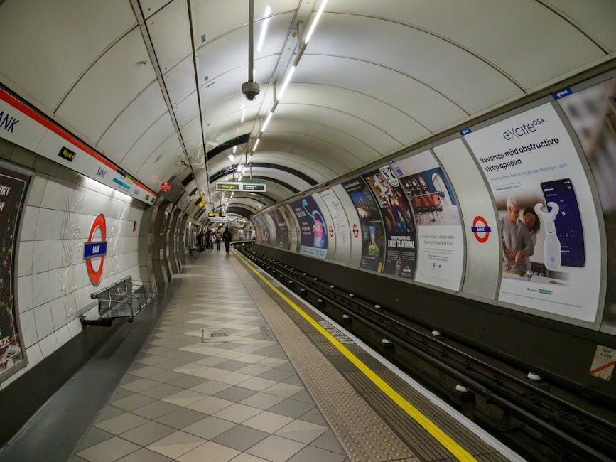 Inside the underground station at Stockwell with a wide curved platform and tiled flooring, featuring the London Underground roundel sign on the tiled wall on the left. A metal bench is positioned near the wall, and a series of illuminated overhead lights run along the ceiling. Advertisements are displayed on the curved wall on the right side, promoting various products and services. The platform is partially occupied by a few commuters walking or standing, some closer to the station’s entrance, with the railway tracks running parallel to the platform edge which has a tactile yellow-strip warning. The scene captures a typical busy underground environment, with a focus on the aesthetics and structure of the station, consistent with the context of house and furniture removals involving transportation through the London transport network, as managed by Man with Van Stockwell.