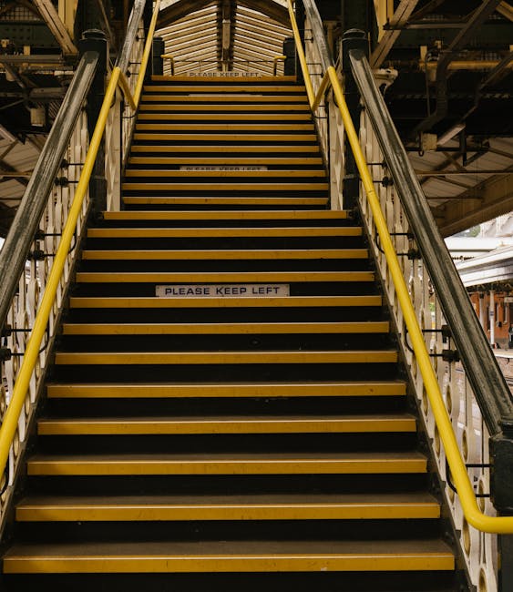 Inside the underground platform at Stockwell station, the image shows a tiled wall with advertisements, including a large Moneybox poster, and directional signage with a yellow arrow indicating the way out. A black metal staircase with orange tiles on the side is located on the left, leading to an upper level. On the platform, there are several cardboard boxes and wrapped furniture items positioned near the edge, ready for a home relocation or furniture transport. A man with van service provider, Man with Van Stockwell, is likely involved in the loading process, which includes carrying and positioning moving boxes and furniture, with a visible section of a red train approaching at the far end of the platform. The lighting is utilitarian, emphasizing the functional environment typical of underground stations, supporting efficient packing and moving logistics for tight access jobs at Stockwell.
